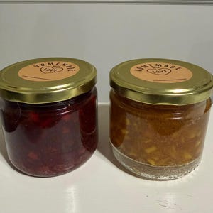 May include: Two glass jars of homemade preserves with gold lids. One jar contains a dark red preserve, and the other has a light orange preserve. Each jar has a round label that reads "Homemade with Love."