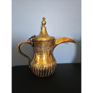 May include: A gold-colored, ornate coffee pot with a curved spout and handle. The pot features intricate etched designs and a decorative finial on the lid. The pot is made of metal and has a vintage appearance.