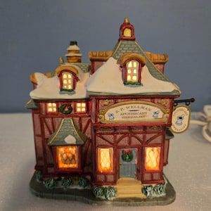 May include: A miniature ceramic apothecary building with a red and brown timber-frame facade, snow-dusted roof, and illuminated windows. A sign above the door reads J.P. WELLMAN APOTHECARY HERBALIST, and a hanging sign features a mortar and pestle icon.