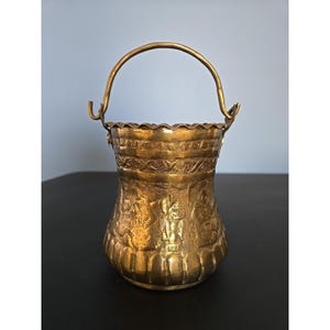 Puede incluir: Un cubo de latón antiguo con un asa curva. El cubo presenta intrincados diseños en relieve alrededor del cuerpo y el borde. El latón tiene una pátina cálida y envejecida, lo que sugiere su naturaleza antigua. El asa está sujeta con soportes curvos.