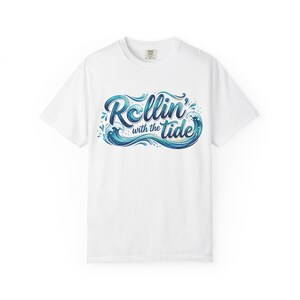 Puede incluir: Camiseta blanca con la frase "Rollin' with the tide" en azul y turquesa, adornada con un diseño de olas. La camiseta tiene cuello redondo y está hecha de un material suave. El gráfico es un diseño estilizado.
