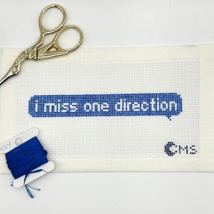 May include: A white needlepoint canvas featuring a blue cross-stitched speech bubble with the text "i miss one direction" in white, accompanied by a small blue "CMS" monogram, gold-toned embroidery scissors, and a spool of blue embroidery floss.