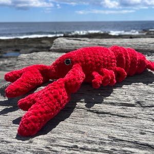 May include: A plush, red lobster toy with black eyes and detailed claws. The soft toy is lying on a weathered wooden surface, with the ocean and sky in the background. A fun, nautical-themed item.