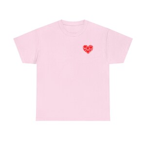 May include: A light pink t-shirt featuring a small heart design made of red hearts on the left chest. The shirt has short sleeves and a crew neck. This casual tee is perfect for everyday wear.