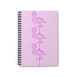May include: A spiral-bound notebook with a light purple cover. The cover features a vertical line of three pink flamingo illustrations. The notebook's spiral binding is black.