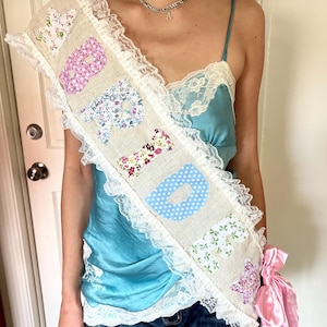 Può includere: Una fascia beige con la scritta "BRIDE" in lettere di tessuto floreale colorato. La fascia è rifinita con pizzo bianco e un fiocco di raso rosa. La fascia è indossata sopra una camisole turchese e jeans blu.