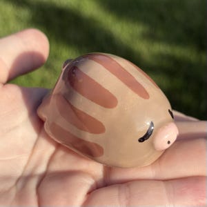 May include: A small, smooth, rounded ceramic figurine shaped like a piglet. The body is a light tan color with darker brown vertical stripes across the back. It features a small, light pink snout with two black dots and closed eyes.