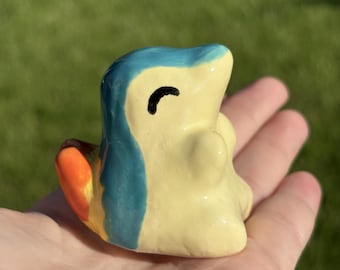 May include: A small, hand-sculpted ceramic figurine shaped like a stylized creature. The figure features a pale yellow body, a blue head and back, and a bright orange tail. It has a simple, hand-painted black curved line representing a closed eye.