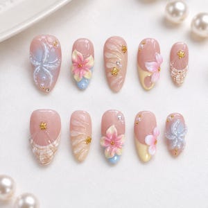 Könnte beinhalten: Ein Set aus zehn künstlichen Fingernägeln mit verschiedenen Designs. Jeder Nagel ist hellrosa und mit floralen und abstrakten Mustern in Pastellfarben wie Blau, Gelb und Weiß verziert. Einige Nägel haben goldene Akzente und Perlen.