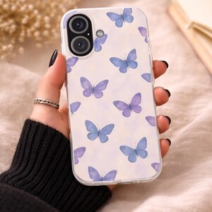 Peut inclure: Coque de téléphone transparente ornée de papillons bleus et violets sur fond blanc. L'étui présente une découpe pour l'appareil photo et est tenu par une personne avec du vernis à ongles noir et une bague argentée.