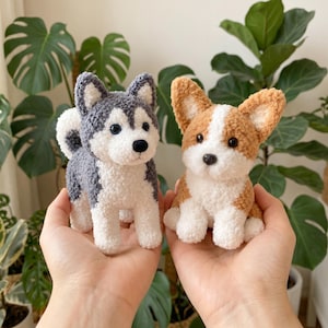 Puede incluir: Dos pequeñas figuras de perro de peluche hechas de hilo texturizado. Uno es un husky gris y blanco con orejas puntiagudas y ojos oscuros, y el otro es un corgi marrón y blanco con orejas grandes y pecho blanco. Ambos tienen narices negras.