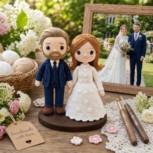 Puede incluir: Adornos para tarta de boda hechos a mano con ganchillo de una novia y un novio. El novio lleva un traje azul marino y la novia un vestido blanco. Una foto enmarcada de una pareja de novios está en el fondo. La etiqueta dice "handmade with love".