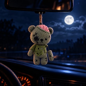 Puede incluir: Un adorno de coche de oso de peluche zombi tejido a ganchillo con cuerpo verde claro, un detalle de cerebro rosa expuesto, un ojo de botón negro y cicatrices cosidas en negro. El oso tiene una camisa verde desgarrada y cuelga de un lazo de cuerda.