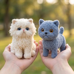 Könnte beinhalten: Zwei kleine, plüschige, gehäkelte Katzenfiguren. Eine Katze ist cremefarben mit einer langen, lockigen Garnstruktur und leuchtend blauen Augen. Die andere Katze ist schiefergrau mit einer kurzen, dichten, flauschigen Struktur und bernsteinfarbenen Augen.