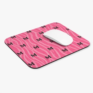 Peut inclure: Tapis de souris rectangulaire avec un motif de rayures zébrées roses et des lettres "M" noires répétées. Une souris sans fil blanche est posée dessus. Le tapis de souris a une bordure noire.