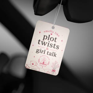May include: A cream-colored tag with the text "smells like plot twists & girl talk" in a whimsical font. The tag features illustrations of a teacup, a book, and flowers in pink. The tag is suspended by a white string.