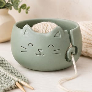 Op de afbeelding: Een saliegroene keramische garenkom in de vorm van een kattengezicht met puntige oren en een gladde, matte afwerking. De kom heeft een spiraalvormige uitsparing aan de zijkant om garen door te voeren en bevat een bol crèmekleurig garen.
