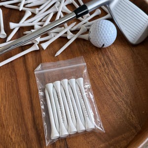 Peut inclure: Image sur le thème du golf avec une balle de golf, un club de golf et de nombreux tees de golf blancs. Un sac en plastique transparent contient un ensemble de tees de golf blancs avec du texte noir. Les articles sont disposés sur une surface en bois.