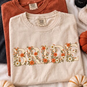 May include: A cream-colored t-shirt featuring the word BRIDE in large, block letters filled with a floral pattern of orange and white daisies and green leaves. The shirt has a Comfort Colors label at the neck and is layered over a rust-colored shirt.
