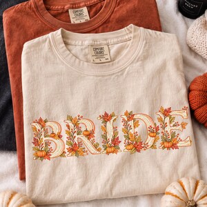 May include: A cream-colored crewneck t-shirt featuring the word BRIDE in large, decorative letters filled with autumn-themed illustrations of orange pumpkins, red and yellow maple leaves, and small berries. The shirt has a Comfort Colors brand label.