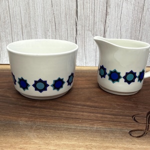 May include: A white ceramic sugar bowl and a matching creamer jug, both featuring a repeating pattern of dark blue and light blue star-shaped geometric motifs around the base. The creamer has a small pouring spout and a side handle.