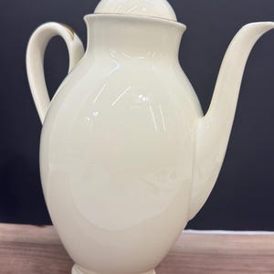 May include: A cream-coloured porcelain coffee pot featuring a smooth, rounded body, a curved handle, and a long spout. The lid is topped with a small, gold-coloured finial, and the base and handle are accented with thin, metallic gold trim details.