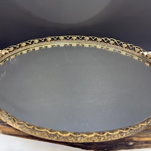 Op de afbeelding: Een ovaalvormig dienblad met een reflecterende spiegelbasis en een decoratieve goudkleurige metalen rand. De rand van het dienblad vertoont een ingewikkeld opengewerkt patroon, aangevuld met twee sierlijke handgrepen aan de zijkanten met metalen details.