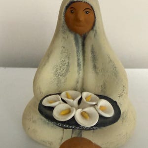 May include: A ceramic figurine of a seated figure wearing a cream-colored hooded garment with a weathered texture. The figure holds a dark oval tray containing several white calla lily flowers with prominent yellow centers in their hands.