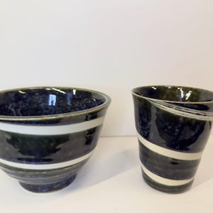 May include: A ceramic bowl and a matching ceramic cup, both featuring a deep navy blue and dark green mottled glaze with prominent horizontal white stripes encircling the exterior of each piece, showcasing a smooth, glossy finish throughout.