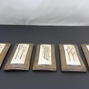Op de afbeelding: Vijf rechthoekige keramische tegels met een donkerbruine rand en een licht crèmekleurig middenpaneel. Elke tegel is voorzien van een unieke donkerbruine lijntekening, waaronder een hagedis, twee hoge planten, een geometrische vorm en bloemmotieven.
