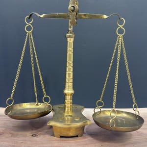 Peut inclure: Une balance en laiton composée d'un montant vertical central et d'un fléau horizontal. Deux plateaux circulaires sont suspendus au fléau par de fines chaînes métalliques. La base présente quatre petites empreintes circulaires.