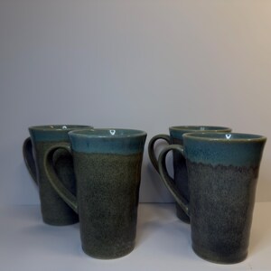 May include: A set of four tall ceramic mugs featuring a two-tone glaze. The upper portion of each mug is finished in a smooth, glossy teal blue, while the lower body displays a textured, speckled dark gray and charcoal finish with a handle.