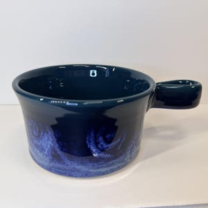 Peut inclure: Un bol à soupe en céramique bleu marine profond avec une large ouverture circulaire et une poignée latérale robuste. L'extérieur présente un motif texturé bleu plus clair près de la base qui s'estompe vers une finition brillante foncée.