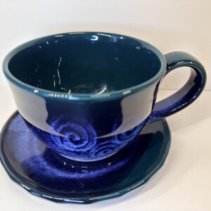 May include: A deep navy blue ceramic teacup and matching saucer set. The cup features a wide, rounded bowl with a sturdy side handle, while the saucer has a slightly wavy, scalloped edge. Both pieces display a glossy, dark blue glaze finish.