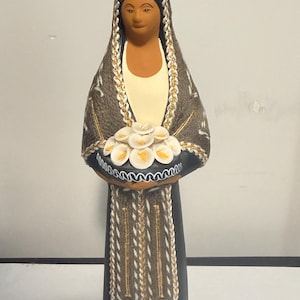 May include: A ceramic figurine of a person wearing a dark gray shawl with white and gold braided trim, holding a bouquet of white calla lilies with yellow centers. The figure has a smooth, matte dark gray base and a light tan complexion.