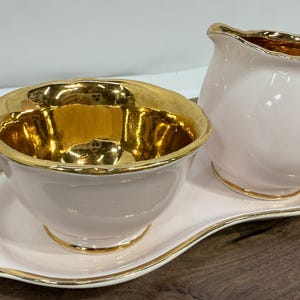 Könnte beinhalten: Ein hellrosa Keramik-Milchkännchen und Zuckerdosenset mit glänzender Oberfläche, metallisch goldener Innenauskleidung und goldfarbenen, gewellten Rändern. Die beiden Teile stehen auf einem passenden hellrosa Ovaltablett mit Goldrand.