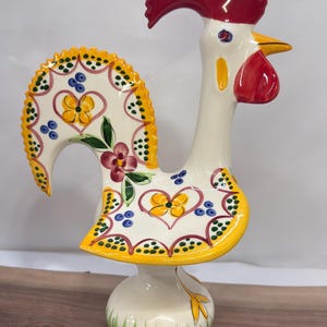 Peut inclure: Une figurine de coq en céramique avec une crête et des barbillons rouge vif, un corps blanc et une queue ainsi qu'une aile bordées de jaune. La surface est ornée de fleurs jaunes, de feuilles vertes et de points bleus peints à la main.