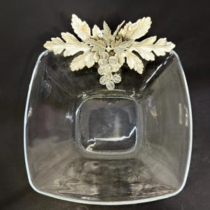 Op de afbeelding: Een vierkante kom van helder glas met een decoratief zilverkleurig metalen accent aan één zijde. Het metalen ornament toont een tros druiven met gedetailleerde bladeren en een klein ovaal label met tekst aan de bovenrand van het blad.