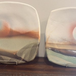 Peut inclure: Deux assiettes carrées en céramique présentant des motifs de paysages à l'aérographe. L'assiette de gauche affiche un soleil circulaire bleu et orange sur des collines. L'assiette de droite montre un soleil pâle au-dessus d'un horizon aux tons terre.