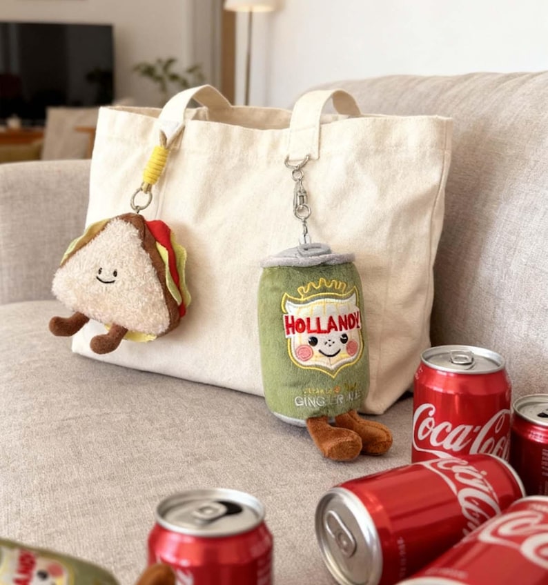 K&ouml;nnte beinhalten: Eine beigefarbene Tragetasche mit zwei Pl&uuml;sch-Schl&uuml;sselanh&auml;ngern: ein l&auml;chelndes Sandwich und eine gr&uuml;ne Limonadendose mit dem Text "HOLLAND!". Mehrere rote Coca-Cola-Dosen sind in der N&auml;he verstreut, was eine spielerische, essensbezogene Szene schafft.