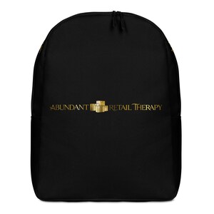 Könnte beinhalten: Ein schwarzer Rucksack mit einem goldfarbenen Logo auf der Vorderseite. Das Logo zeigt den Text ABUNDANT RETAIL THERAPY mit einer kleinen Grafik aus drei gestapelten Kartons in der Mitte. Der Rucksack hat eine abgerundete Oberseite und ein Reißverschlussfach.