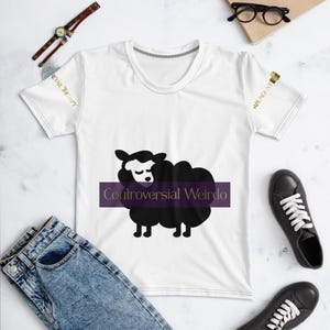 Może przedstawiać: Biały t-shirt z krótkim rękawem z grafiką czarnej owcy wyśrodkowaną na fioletowym poziomym pasku z napisem "Controversial Weirdo" w kolorze złotym. Na prawym rękawie widnieje napis "ABUNDANT", a na lewym "RETAIL THERAPY" w kolorze złotym.