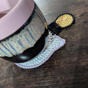 May include: A black and white ceramic mug with a pink rim rests on a handmade crochet coaster. The coaster features a light blue and white body with a lavender border and a yellow and black accent. The mug has a dark, textured design.