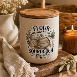 May include: A beige ceramic canister with a wooden lid, displaying a sticker that reads "FLOUR in one hand, SOURDOUGH in the other." The sticker features illustrations of a flour sack, bread, and a bowl. A lit candle and vase are in the background.