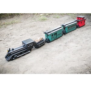 Puede incluir: Un juego de tren de juguete que consta de una locomotora de vapor negra, un vagón de carbón negro con pequeños troncos de madera, un vagón de pasajeros verde y un furgón de cola rojo. Los vagones están conectados en línea con detalles.