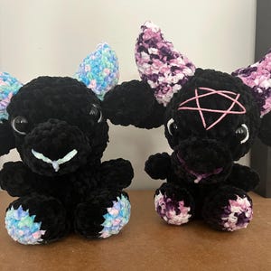 Könnte beinhalten: Zwei schwarze Plüschziegenpuppen mit großen Augen und bunten Hörnern. Eine hat blaue, rosa und grüne Hörner und Hufe, die andere hat lila, rosa und weiße Hörner und Hufe sowie ein rosa Pentagramm. Beide sitzen.