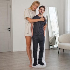 May include: A full-body pillow featuring a printed image of a man in a black t-shirt and dark pants. A woman stands next to the pillow, embracing it. The pillow is white with the man's image printed on it.