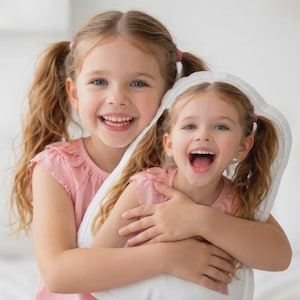Peut inclure: Deux jeunes filles aux cheveux blonds attachés en couettes sourient largement. Elles portent des hauts roses et se serrent dans les bras. L'une d'elles tient un oreiller blanc avec un bord décoratif.