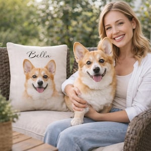 May include: A cushion featuring a corgi portrait with the name "Bella" in script. A live corgi dog with a similar appearance is held by a person. The dog has a tan and white coat and is smiling with its tongue out. The setting is outdoors.