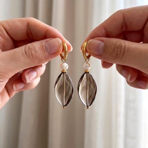 Può includere: Un paio di orecchini a cerchio dorati con un accento di perla e un pendente ovale grigio fumé. Gli orecchini sono presentati su uno sfondo neutro, evidenziando il loro design elegante. Il pendente misura circa 3 cm.